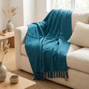 May include: A deep teal throw blanket with a textured, woven fabric finish and knotted fringe along the bottom edge. The blanket is draped over a light beige sofa, showcasing its rectangular shape and consistent solid color throughout.