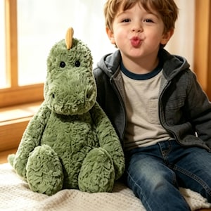 May include: A plush green dinosaur toy with a yellow horn sits next to a child wearing a gray hooded jacket and blue jeans. The dinosaur has a textured, soft appearance. The child is smiling and making a kissing face.