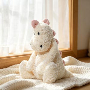 May include: A plush, cream-colored dinosaur toy with pink accents on its ears and back spines. The stuffed animal has black button eyes and is sitting on a cream-colored, textured blanket. The toy is positioned near a window.