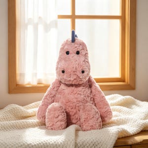 May include: A plush pink hippo stuffed animal with a blue horn. The hippo has black button eyes and a soft, textured body. It is sitting on a cream-colored blanket near a window.