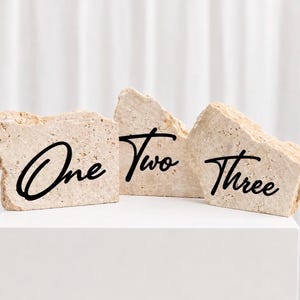Pode incluir: Três blocos de pedra bege com os números "Um", "Dois" e "Três" em escrita preta. Os blocos estão dispostos sobre uma superfície branca contra um fundo branco. Ideal para números de mesa ou decoração.