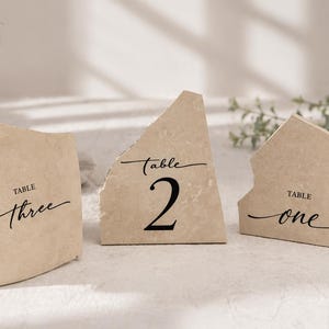 Pode incluir: Três números de mesa de pedra bege com escrita preta. Os números são 1, 2 e 3. As pedras têm uma borda áspera e lascada. Um pequeno vaso de vidro com vegetação está no fundo.