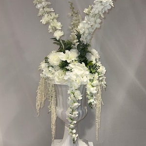 Puede incluir: Arreglo floral blanco en una urna decorativa. El arreglo presenta flores blancas y vegetación, con elementos en cascada. La urna se coloca sobre un pedestal blanco, creando una exhibición clásica y elegante.