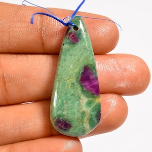 May include: A teardrop-shaped pendant made of polished green zoisite stone with embedded deep magenta ruby inclusions. The stone features a smooth, glossy surface and a single drilled hole at the top, threaded with a thin blue string for suspension.