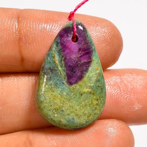 Può includere: Un ciondolo a forma di goccia con una superficie lucida che presenta una prominente sezione centrale viola scuro circondata da texture minerali verde chiaro e verde acqua, con un piccolo foro in alto per un sottile filo rosa.