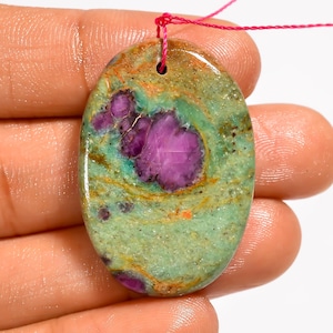 May include: An oval-shaped polished gemstone pendant featuring a smooth surface with a mottled pattern of light green, earthy brown, and vibrant magenta-purple inclusions. A small hole is drilled at the top for suspension with a pink thread.