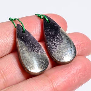 May include: Two teardrop-shaped pendants featuring a dual-tone design with a dark, fibrous purple upper section and a metallic, reflective gold-toned lower section. Each pendant has a small hole at the top with a thin green string attached.