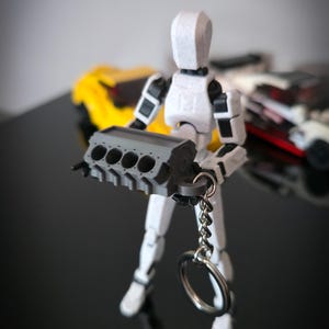 Può includere: Una figura bianca articolata che tiene un portachiavi a forma di blocco motore grigio. La figura ha dettagli neri e si trova su una superficie nera riflettente. Un'auto giocattolo gialla è visibile sullo sfondo.