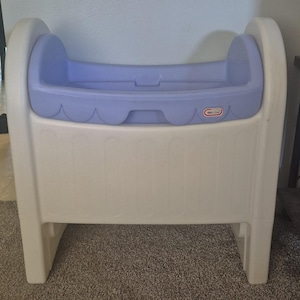 Puede incluir: Una cuna de juguete de plástico con una base de color crema y una sección superior de color morado claro con un borde festoneado. El logotipo de la marca Little Tikes es visible en la parte delantera derecha de la cuna, de diseño redondeado.