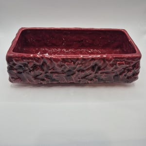 May include: A rectangular ceramic planter with a deep burgundy glaze and a textured, craggy exterior surface. The vessel features a smooth interior basin and a thick, uneven rim, showcasing a glossy finish throughout the entire piece.