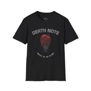 Puede incluir: Camiseta negra con un diseño gráfico de la serie de anime Death Note. El diseño incluye el texto "DEATH NOTE" sobre una manzana roja con un diseño de calavera y el texto "DEATH IS SO CLOSE" debajo.