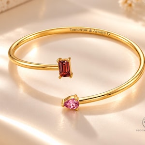 May include: A gold-toned open cuff bracelet featuring a rectangular dark red gemstone and a pear-shaped light pink gemstone at the ends. The inner band is engraved with the text Tomorrow & Always followed by a heart symbol.