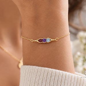 Puede incluir: Una pulsera de cadena dorada con un dije central en forma de vaina de guisante que contiene tres piedras preciosas redondas y facetadas en rojo oscuro, morado y azul claro, unidas a una delicada cadena de eslabones.