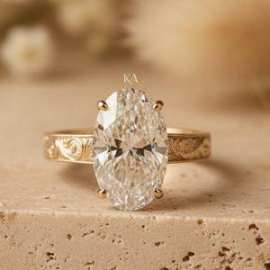 May include: A gold ring featuring a large, oval-cut, clear faceted gemstone held by four prongs. The band is crafted from yellow gold and displays an intricate, engraved floral pattern along its entire outer surface, showcasing fine metalwork.
