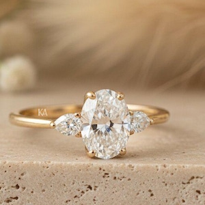 May include: A gold ring featuring a central oval-cut clear gemstone flanked by two smaller pear-cut clear gemstones. The band is thin and polished gold. The letters KA are engraved on the inner side of the band near the setting.