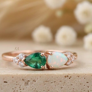 May include: A rose gold ring featuring a pear-shaped green gemstone and a pear-shaped white opal, accented by small round clear stones on both sides. The band is thin and polished, showcasing a delicate design with prong settings for each stone.