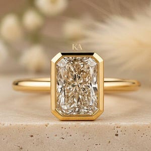 May include: A gold ring featuring a rectangular radiant-cut center stone set in a smooth bezel setting. The band is thin and polished gold. The letters KA are printed in small, delicate font above the center stone of this jewelry piece.