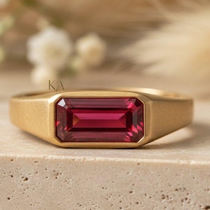 May include: A gold ring featuring a horizontal, emerald-cut, deep pink gemstone set in a matte-finished band. The metal has a smooth, brushed texture, and the stone is rectangular with faceted edges. The letters KA are engraved on the band.