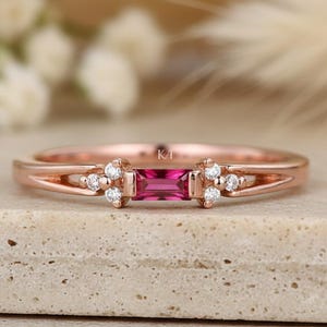 May include: A rose gold ring featuring a central baguette-cut pink gemstone flanked by two clusters of three small round white stones on each side. The band has a split design near the setting and includes the engraved letters K/T inside the band.