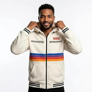 Puede incluir: Sudadera con capucha con cremallera de color crema con un diseño de rayas arcoíris retro en el pecho. La chaqueta presenta rayas negras en las mangas y un pequeño parche con el logotipo "PHM". La sudadera tiene una capucha con cordón y un bolsillo delantero.