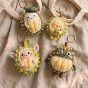 Può includere: Quattro portachiavi a forma di frutto durian fatti a mano all'uncinetto in vari design. Ogni portachiavi presenta un volto sorridente ed è realizzato con filato giallo, verde e marrone. I portachiavi sono esposti vicino a due borse tote all'uncinetto color crema.