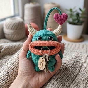 Puede incluir: Un llavero de monstruo de ganchillo turquesa con boca color melocotón, cuernos crema y un corazón rosa. El llavero tiene una etiqueta de madera con la palabra "kolon" y un juego de llaves adjuntas. El monstruo se sostiene en una mano.