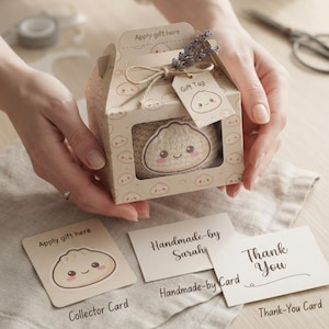 May include: A beige cardboard gift box with a clear window displaying a crocheted dumpling toy. The box features a repeating dumpling pattern, a twine bow, a sprig of lavender, and a gift tag. Includes a collector card and thank-you card.