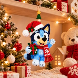 May include: A crocheted blue dog ornament wearing a red and white Santa hat and a red scarf with white snowflake patterns. The dog holds a small green gift box with a red ribbon and is suspended by a string with a small yellow star and tree.
