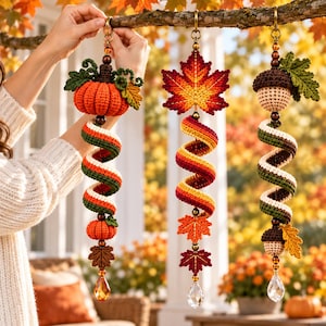May include: Three hanging crochet ornaments featuring autumn themes. One has a pumpkin, one a maple leaf, and one an acorn. Each includes a spiral tail in orange, green, and cream yarn, accented with wooden beads and a clear crystal drop.