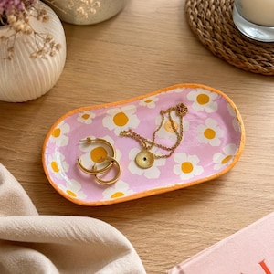 Peut inclure: Un vide-poche ovale en céramique avec une base rose clair, orné de fleurs marguerites blanches et jaunes et d'un bord orange vif. Le plateau contient une paire de créoles dorées et un collier à chaîne dorée avec un pendentif circulaire.
