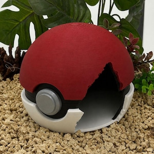 Può includere: Una struttura sferica e cava in plastica rossa e bianca che ricorda una Poké Ball. La metà superiore è rossa, quella inferiore è bianca e una fascia nera con un bottone circolare grigio centrale circonda il centro. È visibile un'ampia apertura.