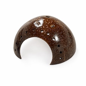 May include: A dark brown, polished coconut shell dome featuring a smooth, curved surface with a series of small, circular holes drilled along the edges and top. The natural fibrous texture of the coconut shell is visible throughout the exterior.