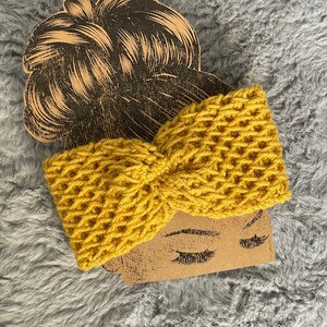 May include: A mustard yellow knitted headband with a textured waffle stitch pattern. The headband is displayed on a brown cardboard cutout of a woman's head and face, with closed eyes and long eyelashes. The background is a grey, fuzzy fabric.