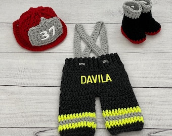 May include: A crocheted firefighter outfit consisting of a red hat with a grey front patch displaying the number 37, black overalls with grey suspenders and yellow reflective stripes, and a pair of black and grey crocheted boots with the name DAVILA.