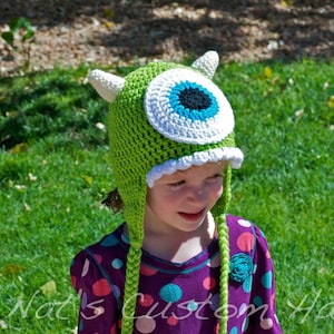 May include: A green crocheted hat with a large, white-rimmed eye and two white horns. The hat has a white scalloped brim and green braided ties. The hat is worn by a person wearing a purple polka dot shirt. The text "© Nat's Custom Hats" is visible.