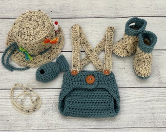 Puede incluir: Un conjunto de bebé tejido a crochet con temática de pescador. El conjunto incluye un gorro, un mono, botines y un juguete de pez. El gorro es beige con un borde marrón y un señuelo de pesca rojo. El mono es azul con tirantes beige y un botón marrón. Los botines son beige con un ribete marrón. El juguete de pez es azul con un ojo blanco.