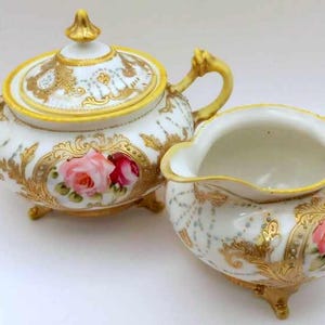 May include: A white porcelain sugar bowl and creamer set with gold trim and floral designs. The set features pink and red roses with green leaves.