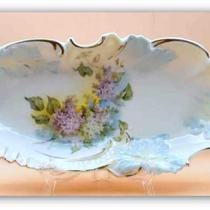 May include: An oval, antique porcelain serving dish with a ruffled edge and gold trim. The dish features a hand-painted floral design with purple, yellow, and green flowers. The dish is displayed on a clear stand.