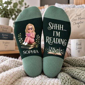 May include: A pair of dark green socks with light green cuffs. The left sock features a cartoon illustration of a person with blonde hair and glasses reading a book, with the name SOPHIA printed below. The right sock displays the text SHHH... I'M READING above an open book graphic.