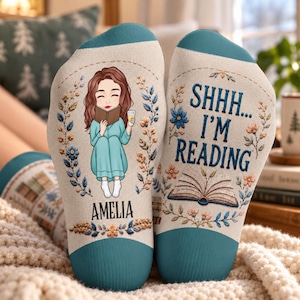 May include: A pair of cream-colored socks with teal cuffs. One sock features an illustration of a person reading a book with the name AMELIA printed below. The other sock displays the text SHHH... I'M READING above an open book graphic.