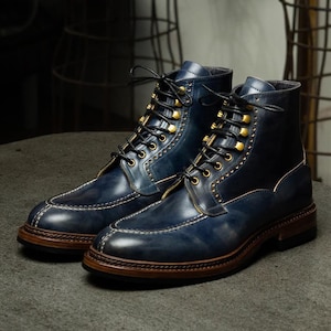 May include: A pair of navy blue leather boots featuring a moc-toe design with white contrast stitching, black laces, and gold-toned eyelets. The boots have a dark brown stacked leather midsole and a sturdy outsole, showcasing a polished finish.