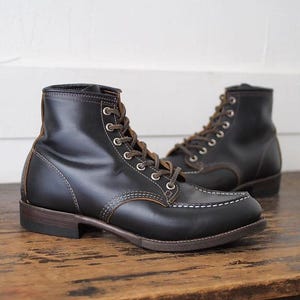 Puede incluir: Un par de botas de cuero negro con puntera estilo mocasín, costuras blancas en contraste, ojales metálicos plateados y cordones de color marrón oscuro. Las botas tienen un tacón apilado y una suela de color marrón oscuro.