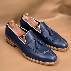 Pode incluir: Um par de mocassins de couro azul-marinho com uma borla no peito do pé, debrum branco ao longo do colarinho e um salto e sola de cor castanho-claro. Cada sapato contém uma forma de madeira com um botão dourado no calcanhar.
