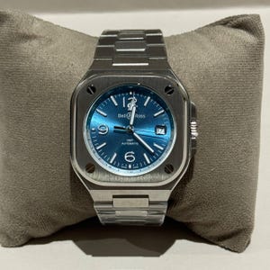 Op de afbeelding: Een roestvrijstalen Bell & Ross horloge met een vierkante wijzerplaat en een blauwe wijzerplaat. Het horloge heeft een zilveren band en wordt gepresenteerd op een grijs kussen. De wijzerplaat heeft witte cijfers en wijzers, met de merknaam en "GMT AUTOMATIC".
