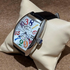 Könnte beinhalten: Eine silberfarbene Armbanduhr mit schwarzem Lederarmband. Das Zifferblatt ist weiß mit bunten, übergroßen Ziffern und der Aufschrift "FRANCK MULLER GENEVE". Die Uhr liegt auf einem beigen Kissen.