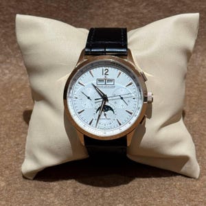 May include: A rose gold wristwatch with a white face, black leather strap, and multiple sub-dials. The watch displays the date and moon phase. The watch is displayed on a beige cushion.