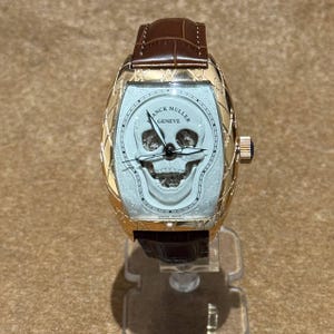 May include: A gold-toned wristwatch with a brown leather strap. The watch face has a skull design and the words "FRANCK MULLER GENEVE". The watch is displayed on a clear stand.