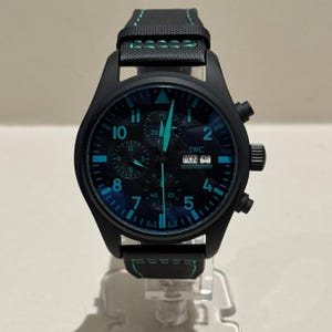 Puede incluir: Reloj IWC Schaffhausen negro con correa negra y detalles turquesa. La esfera muestra el día de la semana y tiene números y agujas turquesas. El reloj tiene función de cronógrafo y una correa texturizada con costuras turquesas.