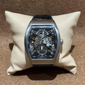 Op de afbeelding: Een zilverkleurig Franck Muller Vanguard horloge met een zwarte leren band, liggend op een beige kussen. De wijzerplaat toont ingewikkelde interne mechanismen en de tekst "VANGUARD NORTH" en "SOUTH SEVEN DAYS".
