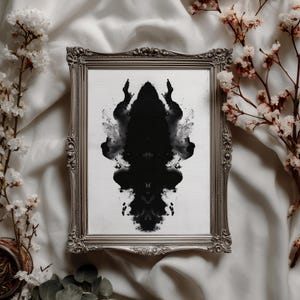 May include: A rectangular silver ornate frame containing a white print with a symmetrical black inkblot design. The inkblot features dark, fluid, and abstract shapes that mirror each other vertically, creating a Rorschach-style pattern.